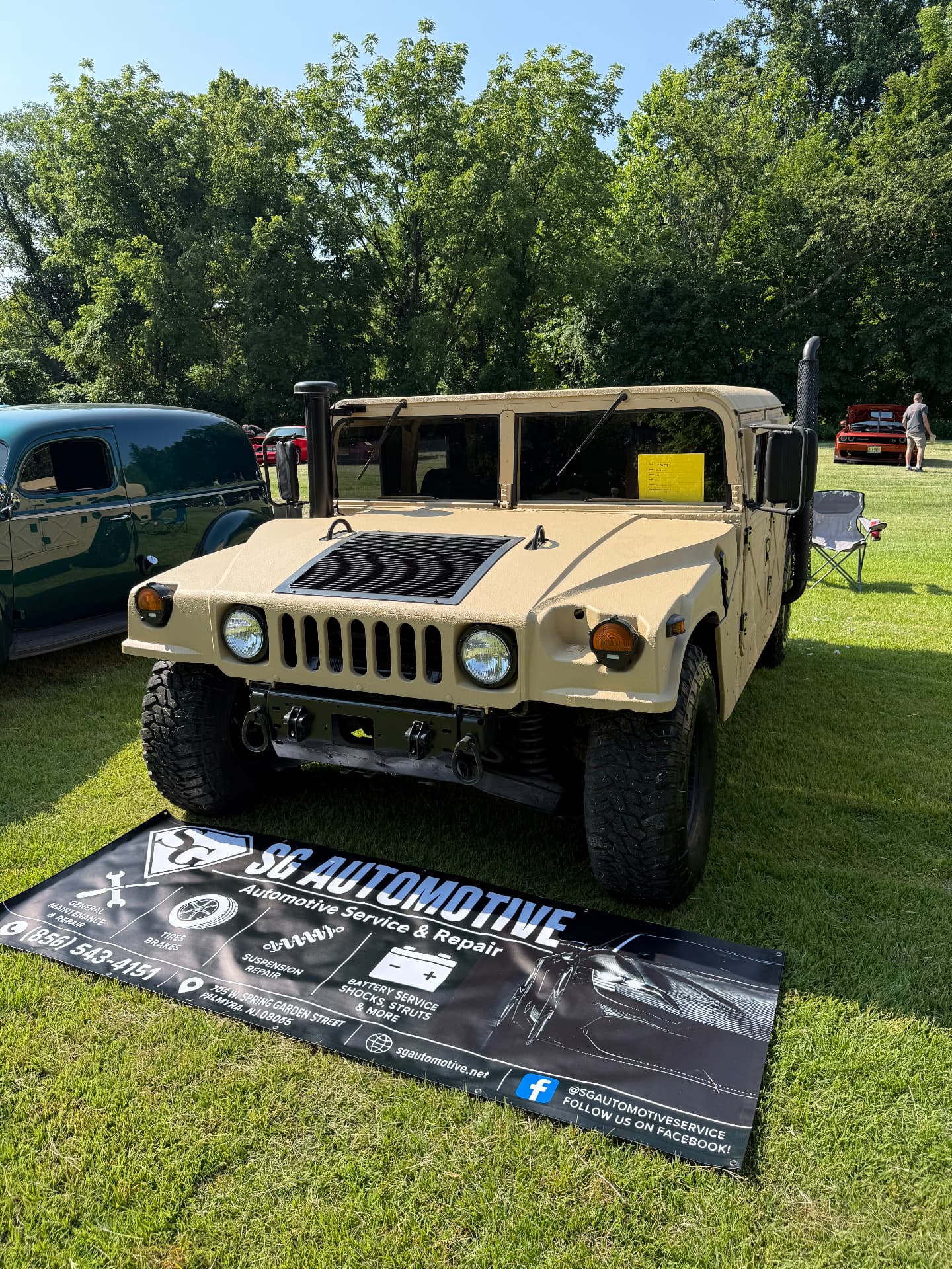 Tan military Humvee on display beside the SG Automotive Service & Repair banner at a car show