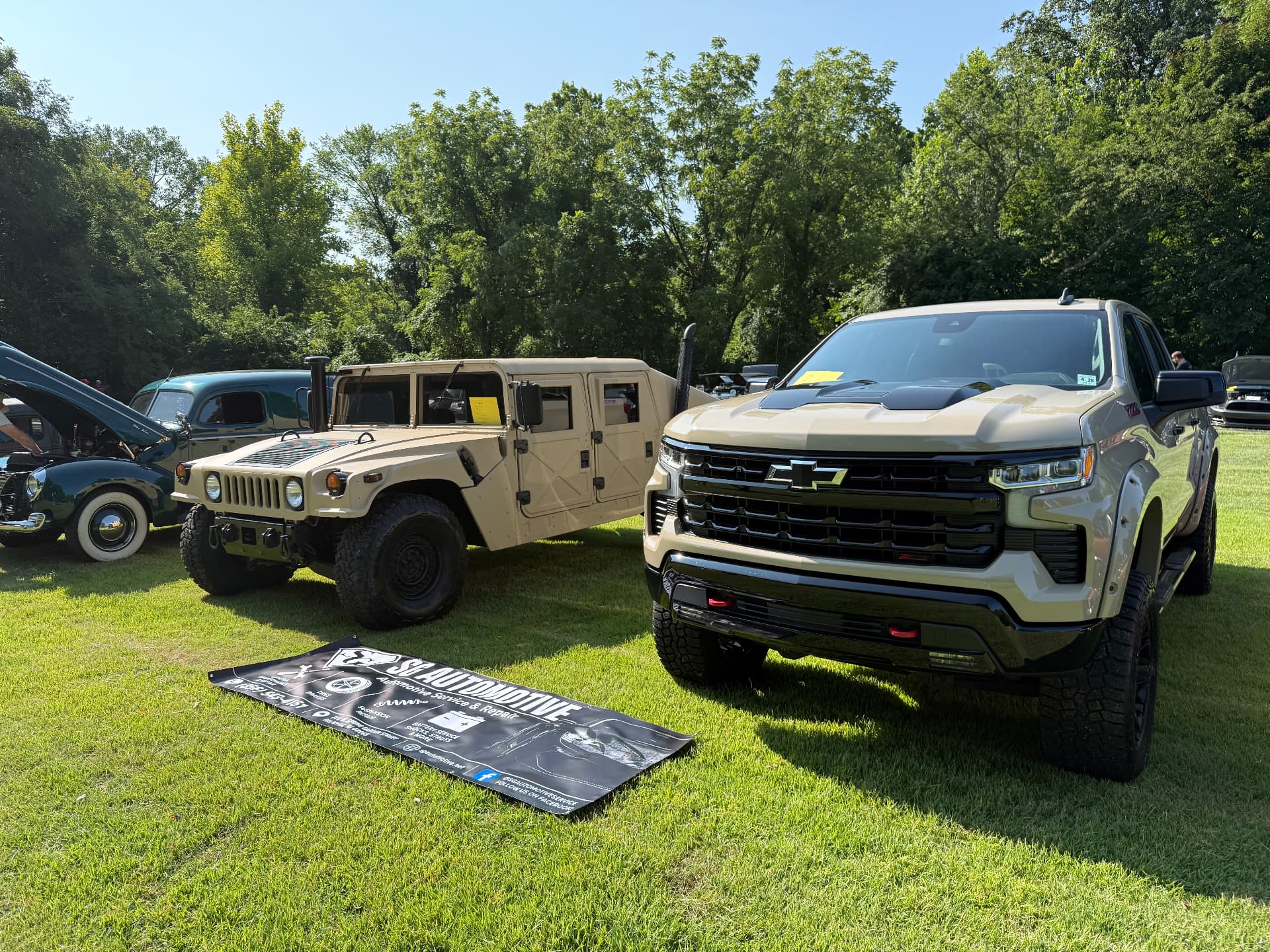 Tan Humvee H1 and matching Chevrolet Silverado Trail Boss at a local car show with SG Automotive banner